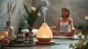 Ein Diffusor gibt Dampf auf eine Holzmatte mit Flaschen mit ätherischen Ölen ab, während im unscharfen Hintergrund eine Frau meditiert und ihr emotionales Wohlbefinden durch Aromatherapie steigert.